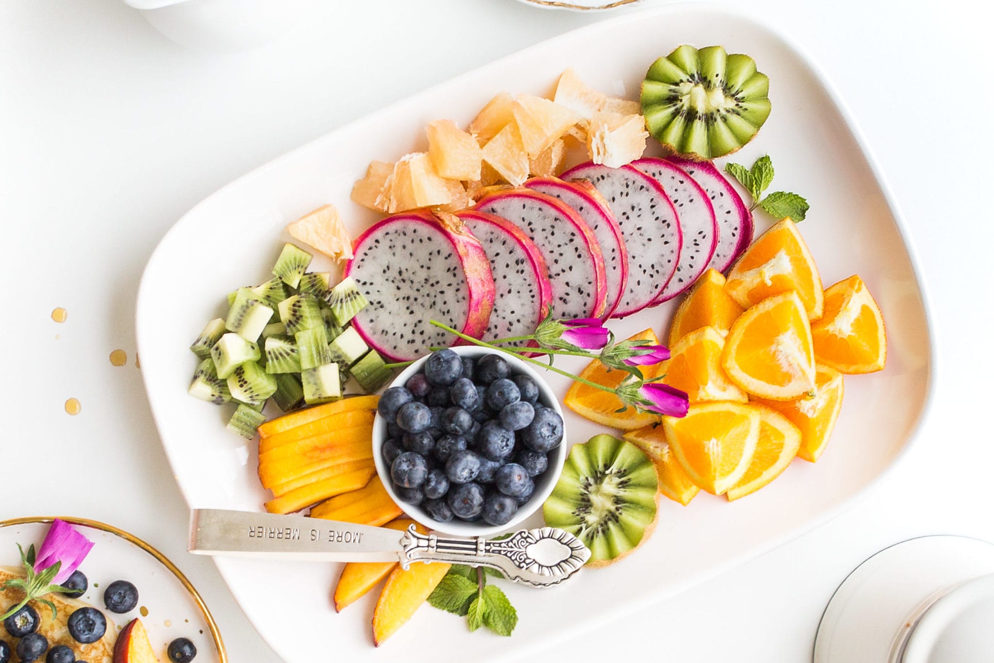 A vibrant fruit platter with sliced kiwi, dragon fruit, oranges, and blueberries, garnished with mint leaves on a white background.
