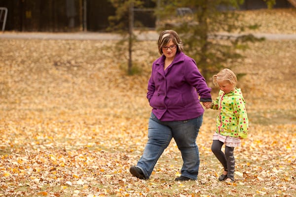 A woman in a purple jacket walks hand-in-hand with a young girl in a green coat through a park covered in autumn leaves.