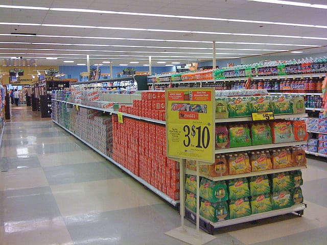 Aisle in a supermarket displaying various beverages and snacks on shelves, with price tags visible under fluorescent lighting.