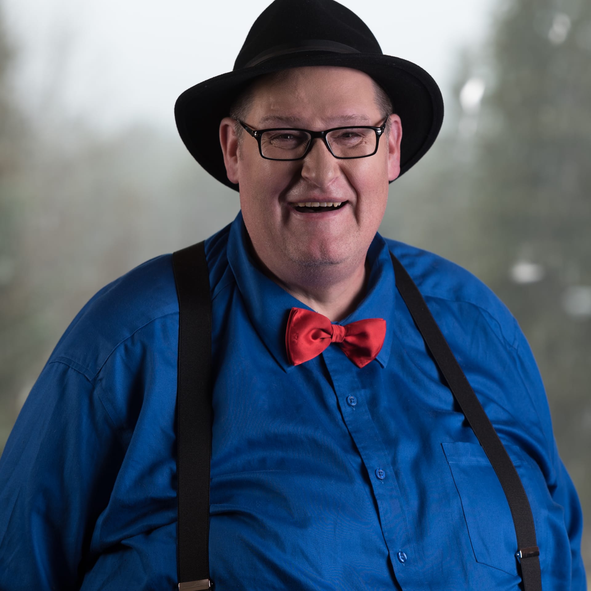 A cheerful man wearing a black hat, blue shirt, red bow tie, and suspenders, smiling at the camera with a blurred background.