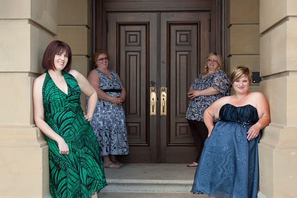 Four women smiling in front of a large wooden door, each dressed in formal wear, with the first woman standing prominently in a green dress.