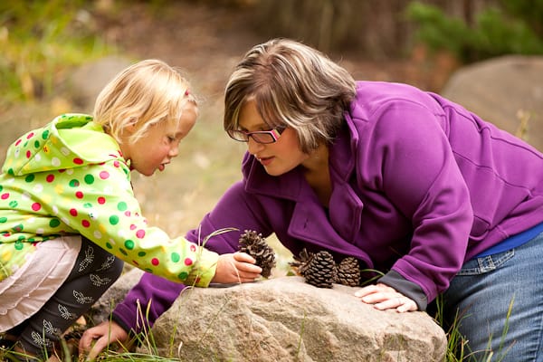 A young girl and a woman examining pine cones together in a forest setting.