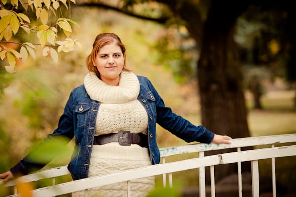 A woman in a white sweater and blue denim jacket leans on a white railing outdoors, surrounded by autumn leaves.