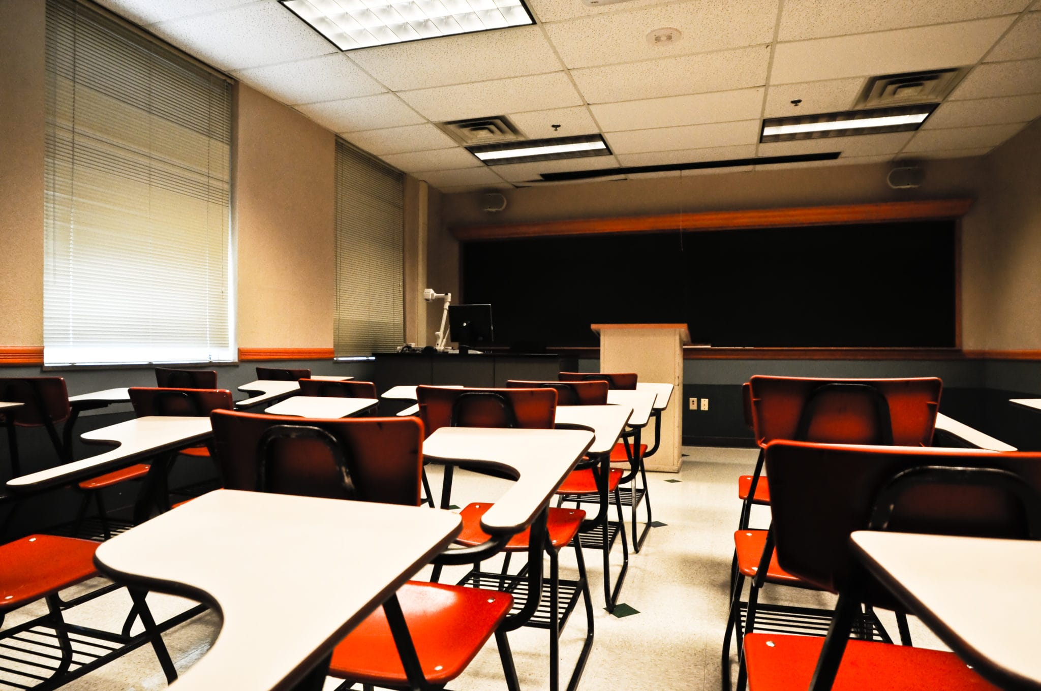 Empty classroom with rows of desks and chairs facing a blackboard, under bright artificial lighting.