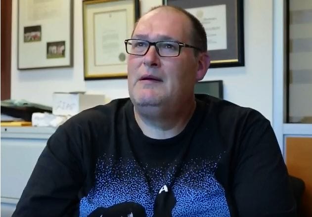 Middle-aged man wearing glasses and a black shirt with a speckled pattern, sitting in an office surrounded by framed documents.