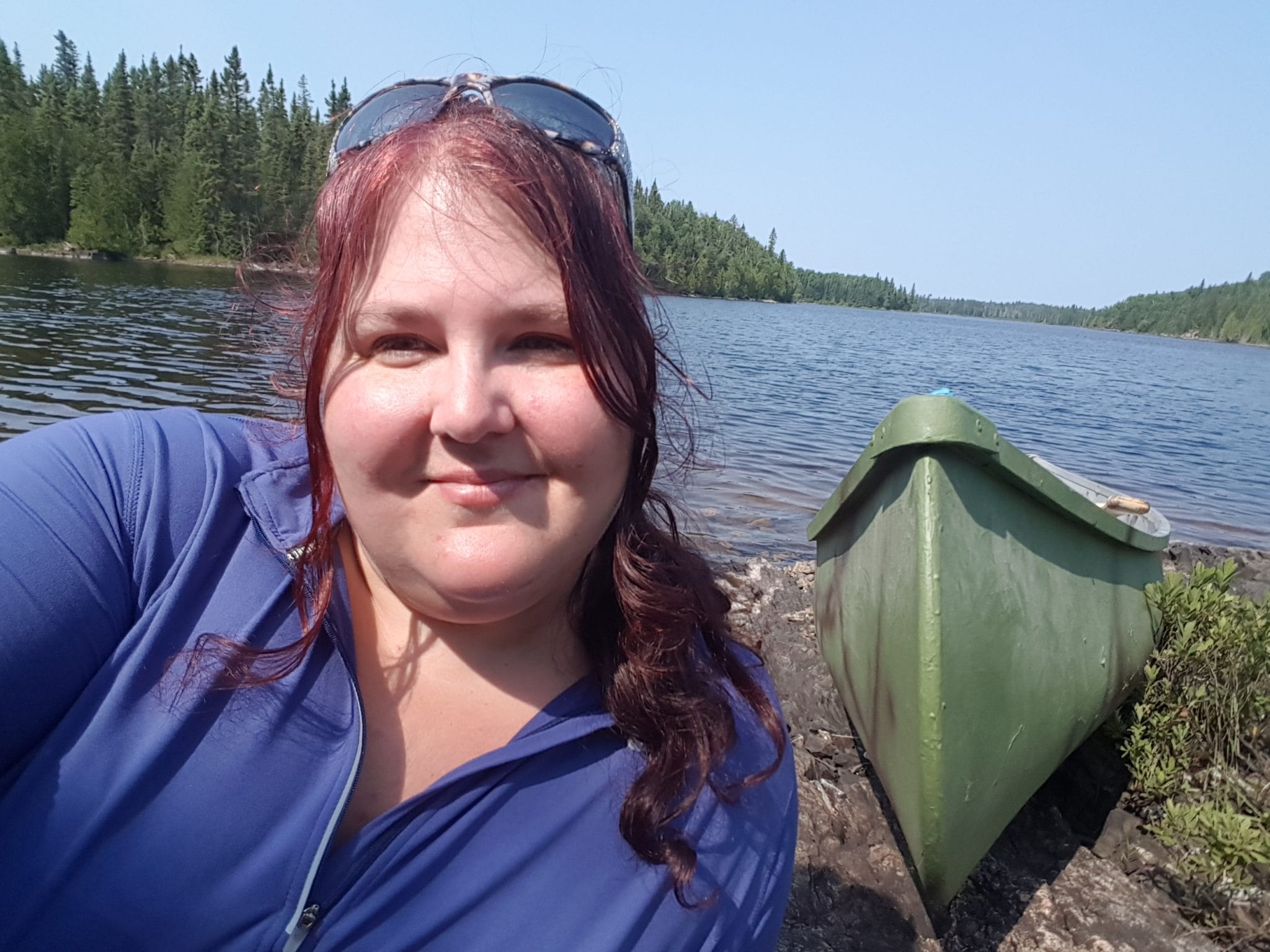 A woman with red hair and sunglasses on her head smiling at the camera, by a lake with a green canoe nearby.