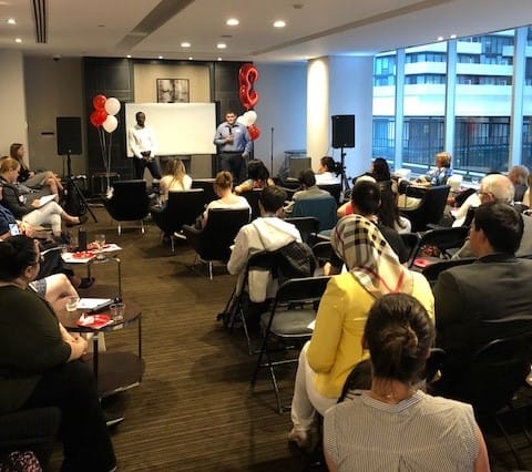 A corporate event with two speakers addressing an audience seated in a room decorated with red and white balloons.
