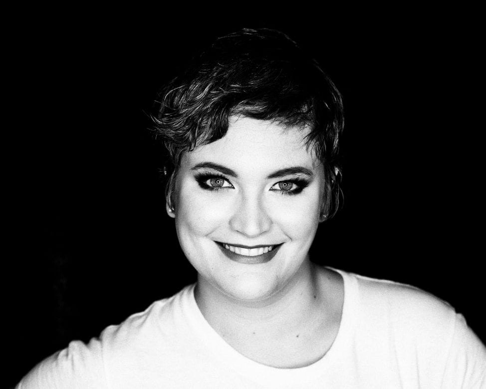 Black and white portrait of a smiling woman with short hair, wearing a white top against a dark background.
