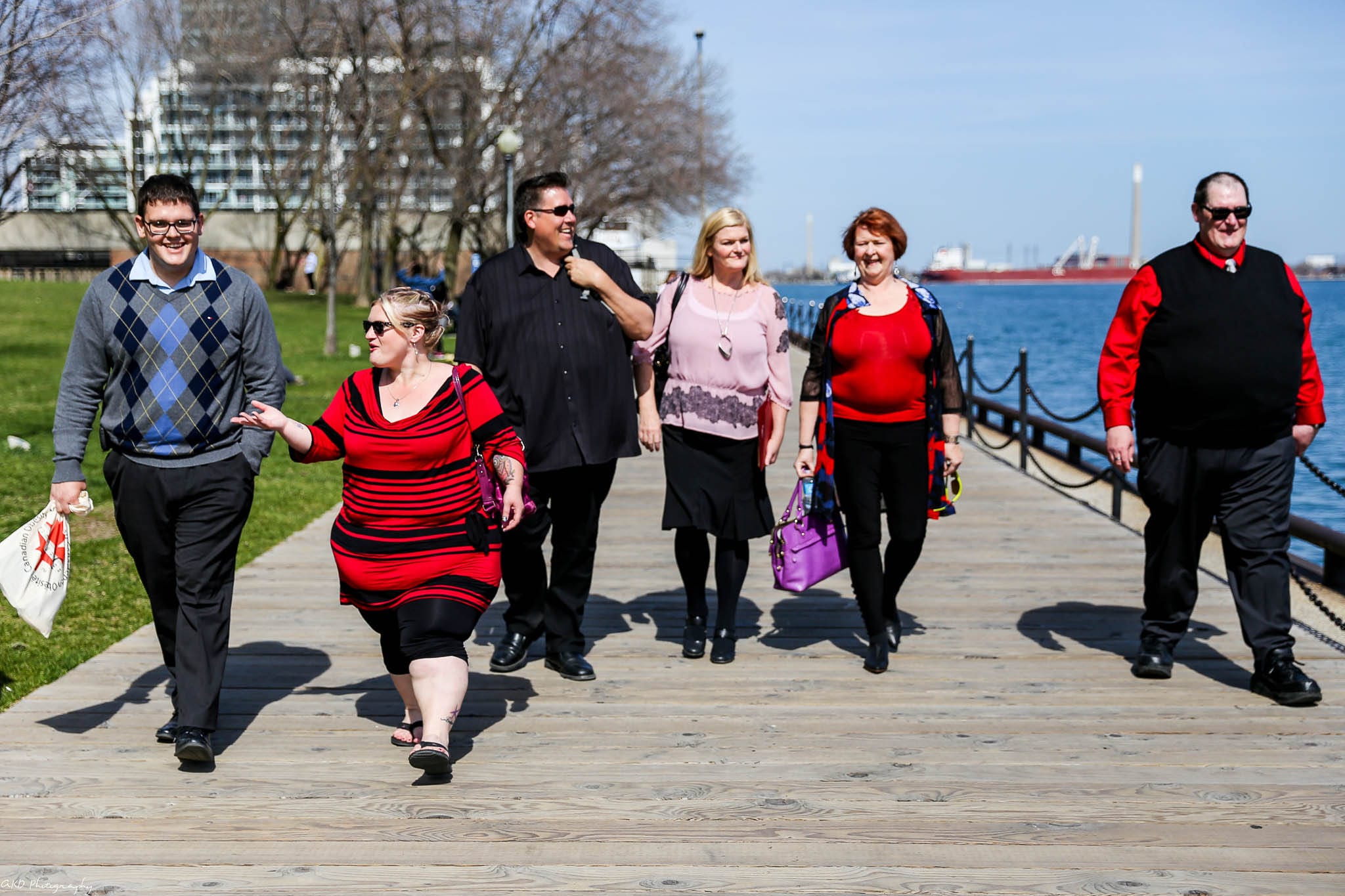 A group of six adults walking together on a wooden boardwalk by a lakeside, carrying shopping bags and smiling on a sunny day.