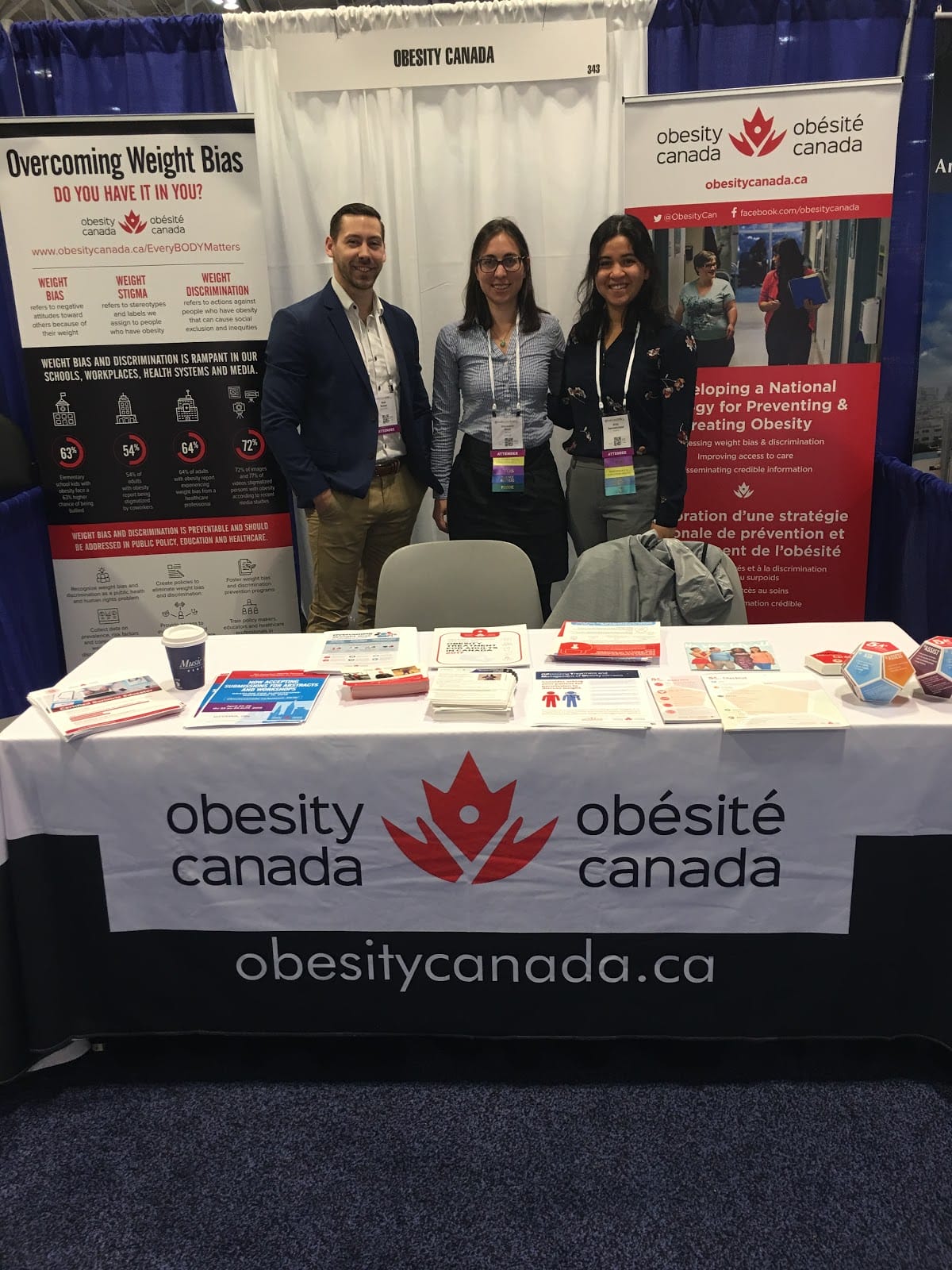 Three people stand behind an obesity canada informational booth at a conference, smiling, surrounded by educational materials and banners.