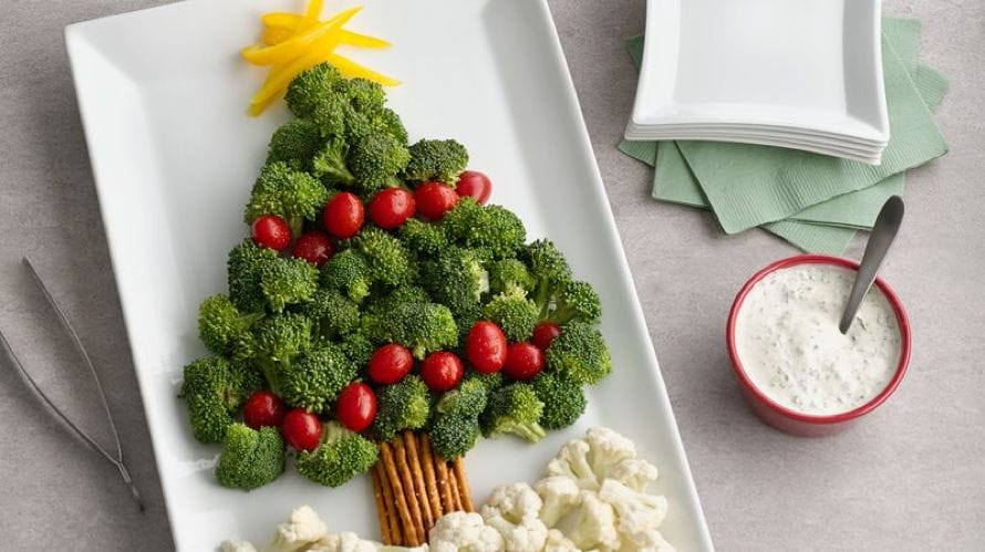 A festive broccoli tree with cherry tomato decorations on a plate, accompanied by cauliflower, pretzel sticks, a dip bowl, and napkins.