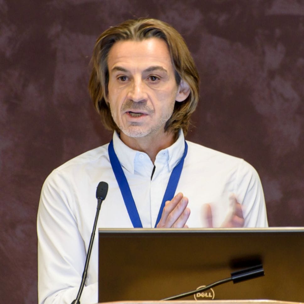 Man with long hair presenting at a podium with a laptop and wearing a name badge and white shirt.