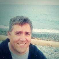Man smiling at the camera with a pebbly beach and ocean in the background.