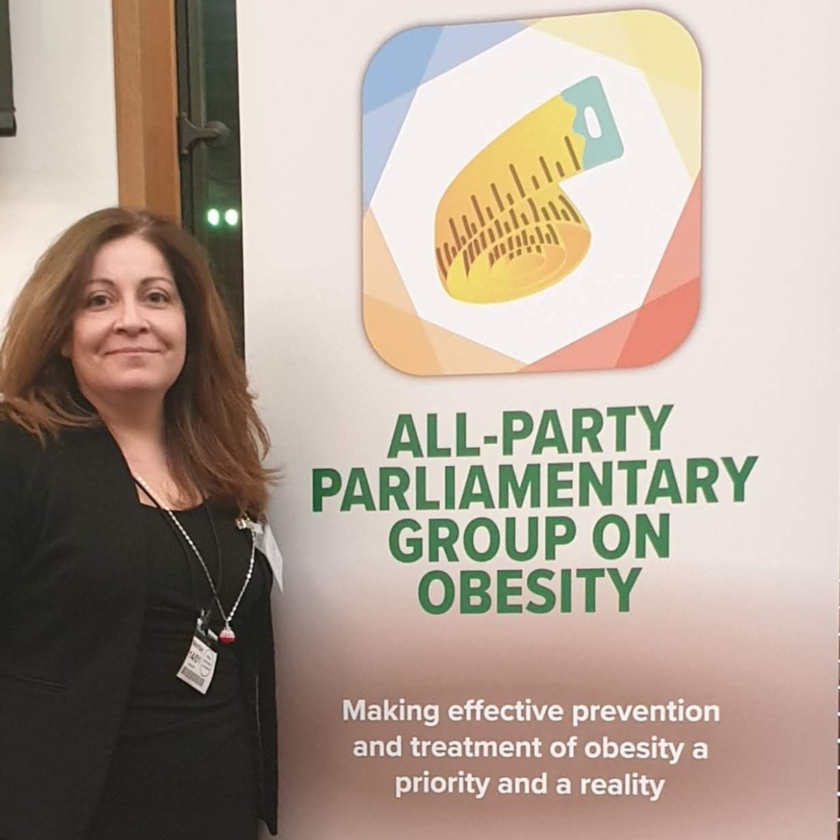 A woman standing beside a banner of the all-party parliamentary group on obesity, featuring its logo.