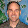 A smiling man with a map background, wearing a gray shirt.