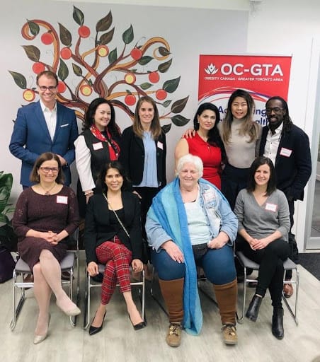 Group of nine diverse people posing for a photo in a room with a large tree mural on the wall, featuring the logo of obesity canada.
