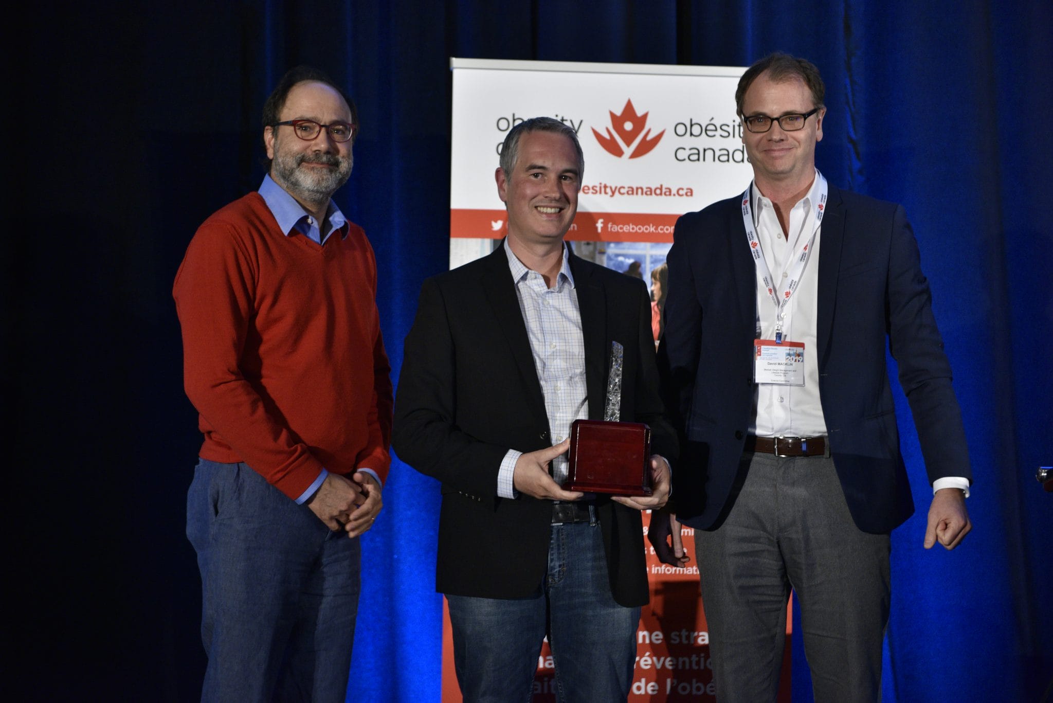 Three men on a stage at an event, one receiving an award, with a banner for obesity canada in the background.