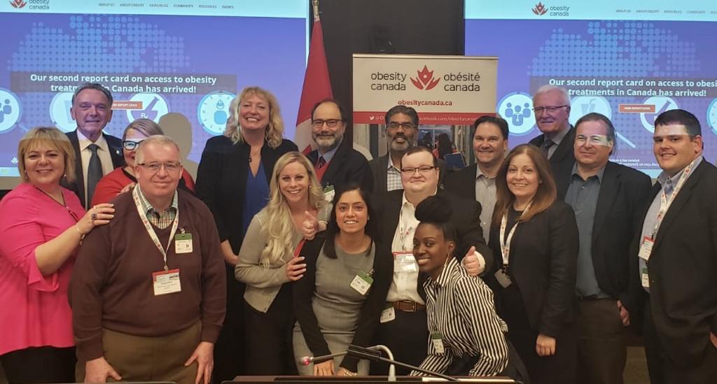 A group of professionals smiles for a photo at an obesity canada event, standing in front of a banner displaying the organization's logo.