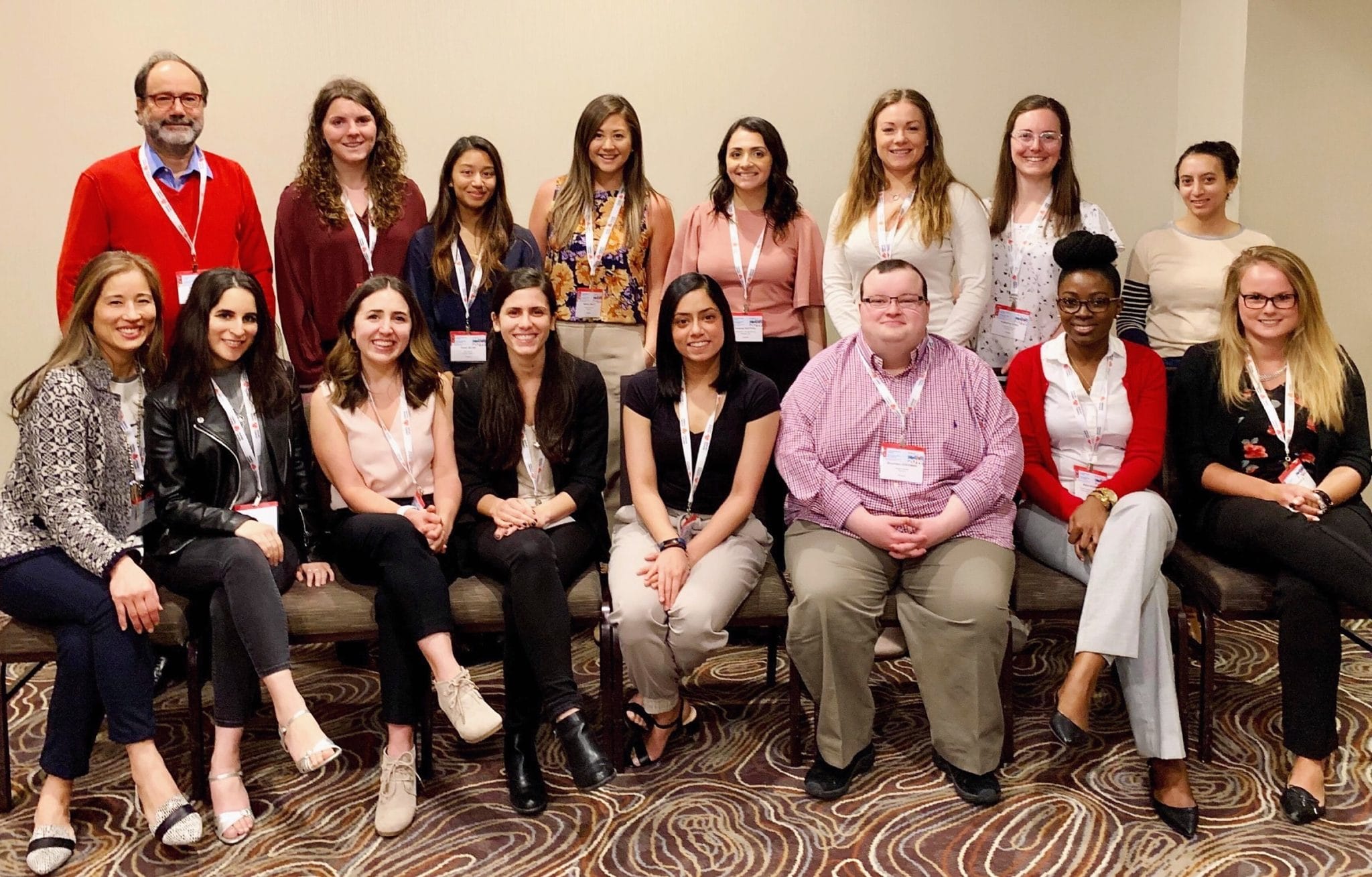 Group of sixteen diverse people posing for a photo at a conference, some sitting and others standing in a hotel meeting room.
