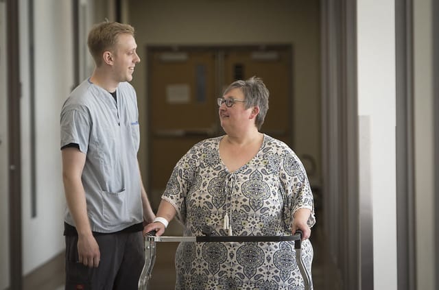 A nurse assisting a woman using a walker in a hospital corridor, both are conversing and smiling.