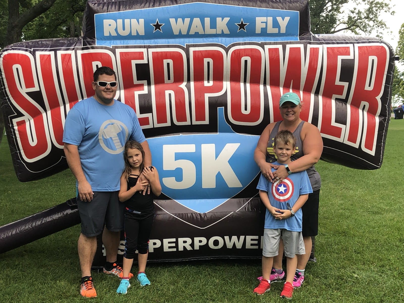 Family of four posing in front of a "superpower 5k" race banner at an outdoor event, with two children holding superhero shields.