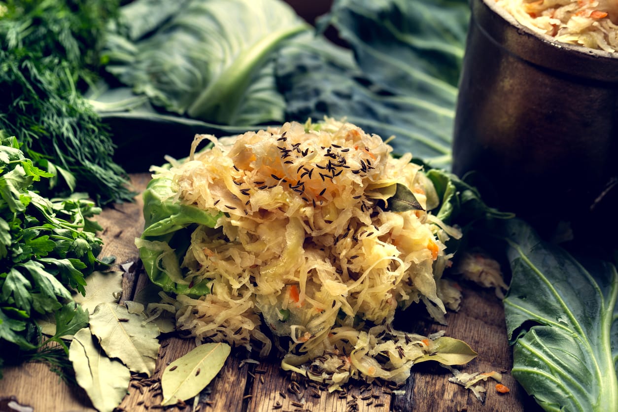 A wooden bowl filled with fresh sauerkraut topped with caraway seeds, surrounded by green vegetables and herbs on a rustic table.