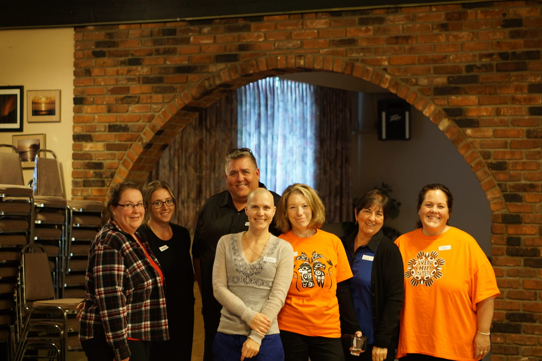 Group of seven adults smiling in a room with a brick arch, three in orange shirts and two wearing name tags.