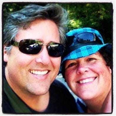 A close-up selfie of a smiling man and woman wearing sunglasses and a hat, outdoors on a sunny day.