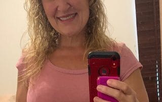 A woman with curly hair smiles while taking a selfie with a red phone in a mirror, wearing a pink t-shirt.