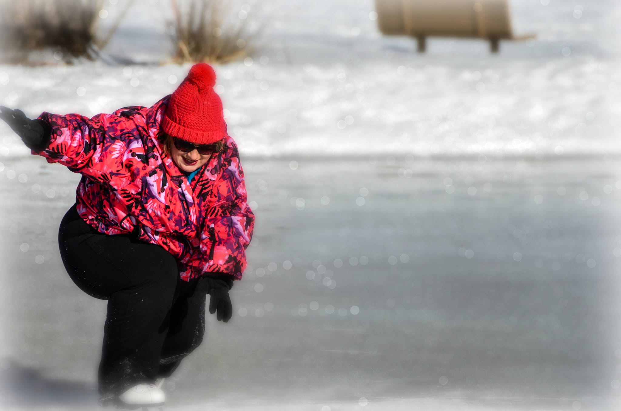 A woman in a vibrant pink jacket and red beanie skates gracefully on an outdoor ice rink, her posture focused and dynamic.