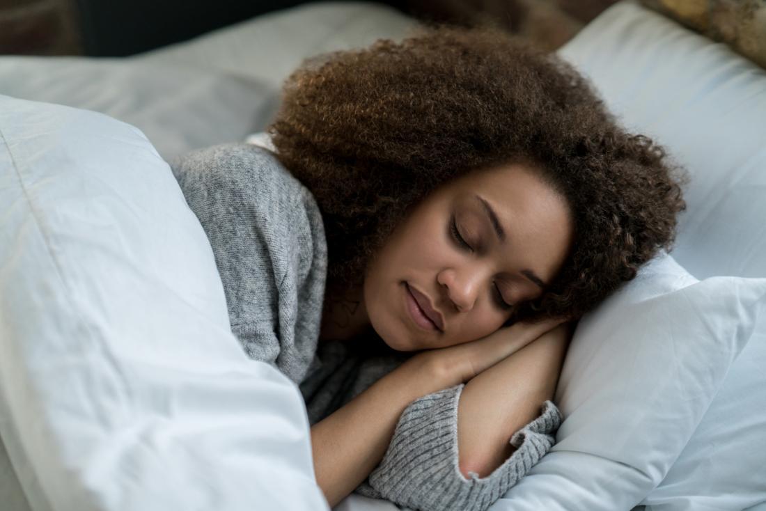 A woman with curly hair peacefully sleeping in a bed, wrapped in a white comforter and wearing a gray sweater.