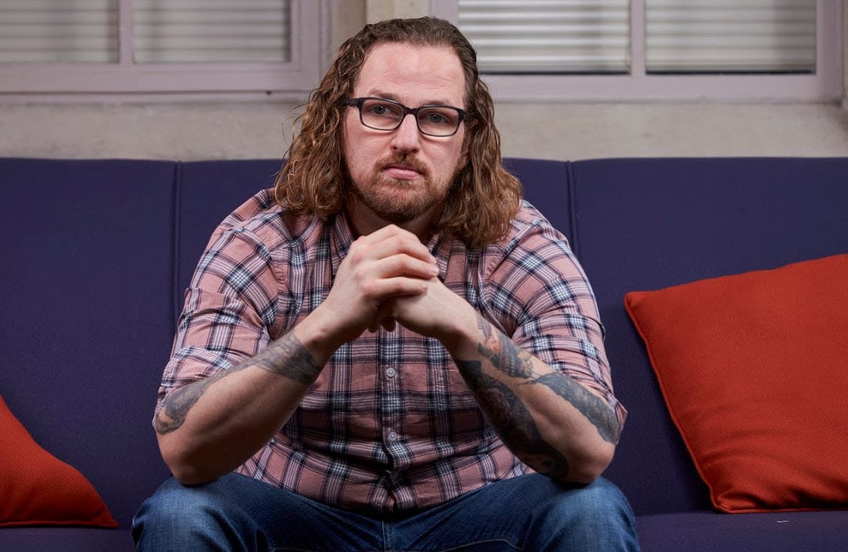 A man with long curly hair and glasses wearing a plaid shirt sits on a purple sofa, looking directly at the camera with a serious expression.