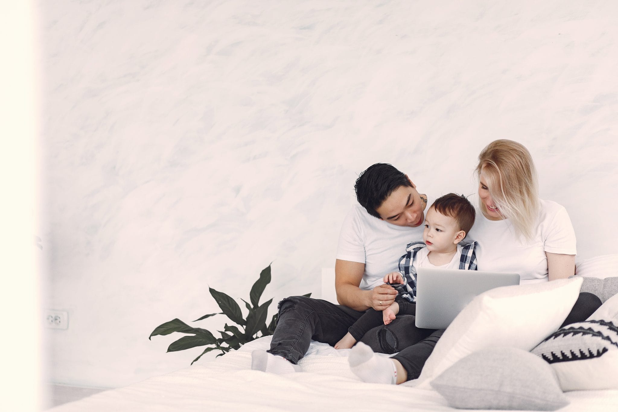 A family sits on a bed; a man and a woman lovingly look at a child who clutches a toy, with a laptop open in front of them.