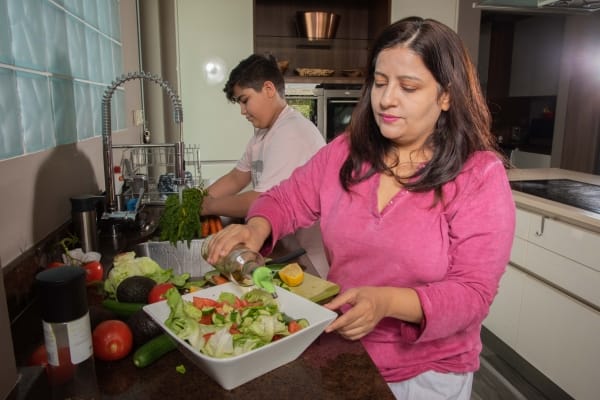 A woman in a pink shirt prepares a salad in a kitchen, pouring dressing, while a young man in the background washes vegetables at the sink.