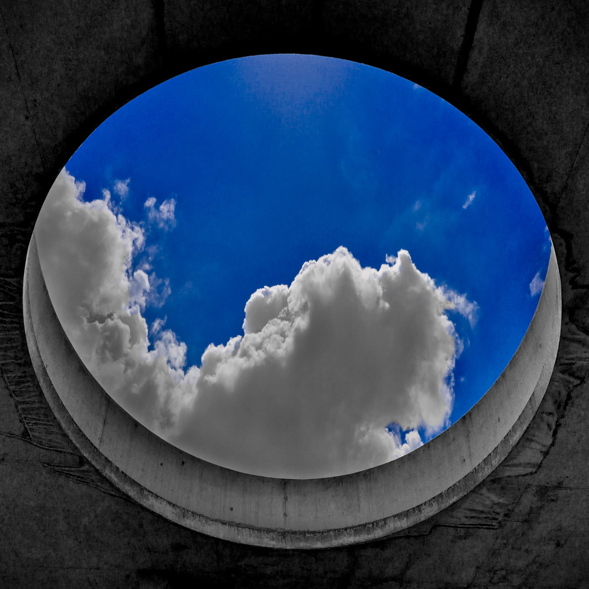 View of a bright blue sky with fluffy clouds framed by a circular concrete structure, emphasizing a stark contrast between the natural and built environments.