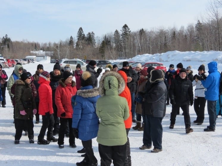 Group of people wearing winter clothing gathered in a snowy parking lot, some are talking while others look on.