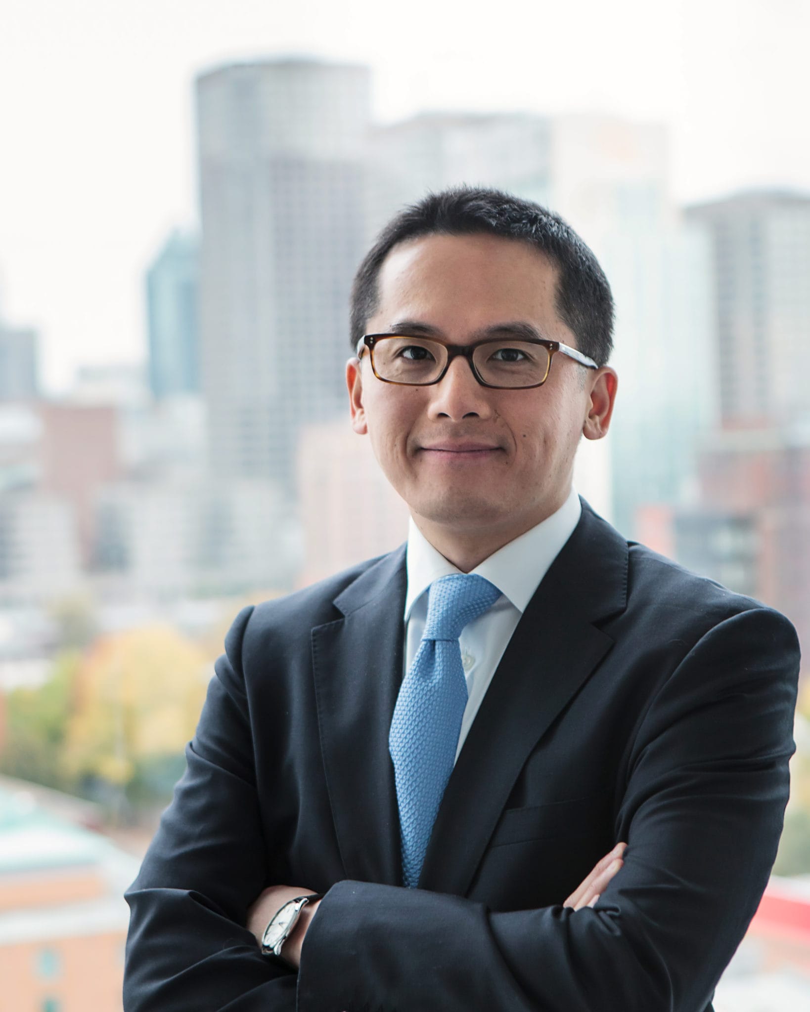 A professional asian man in a blue suit and glasses smiling at the camera with a city skyline in the background.