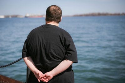 Man in a black shirt standing by a waterfront, looking out over blue water with ships in the distance, holding his hands together behind his back.