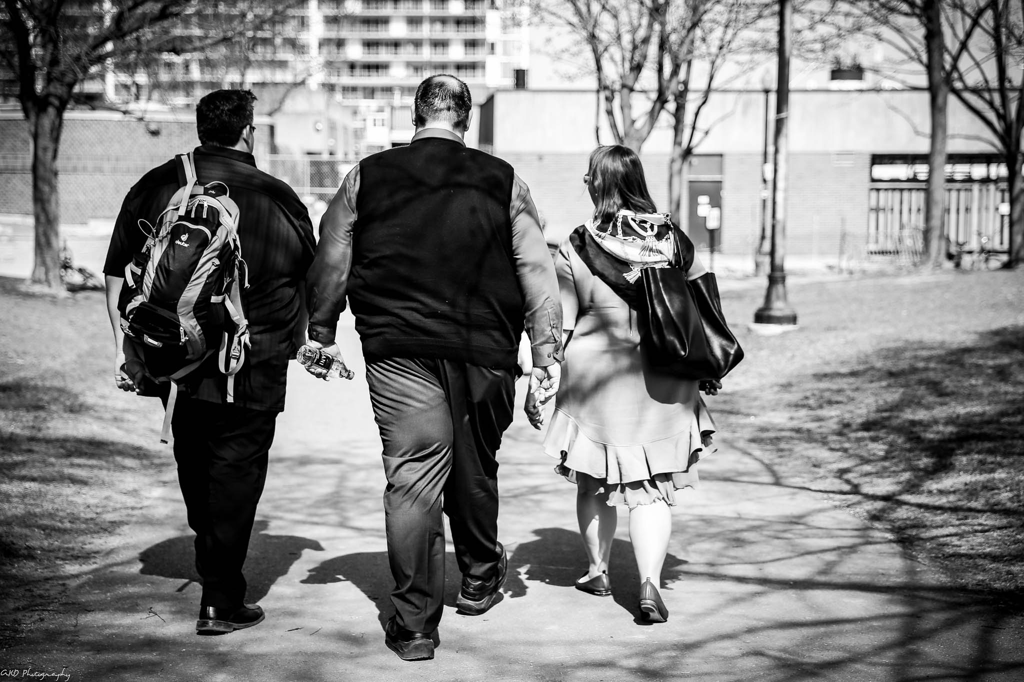 Three people walking in a park, viewed from behind. two men in suits and one woman in a skirt, all carrying bags.