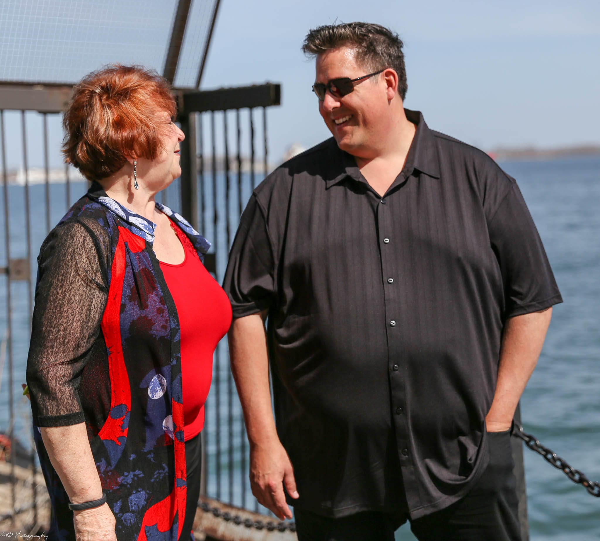 A man and a woman, both smiling and looking at each other, standing by a waterfront with a barrier and the sea in the background. the woman is dressed in a red and blue dress, and the man in a black shirt.