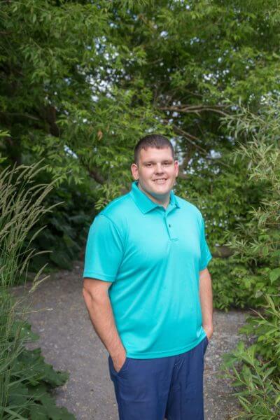 A man in a turquoise polo shirt smiling, standing on a garden path surrounded by green bushes and tall grasses.