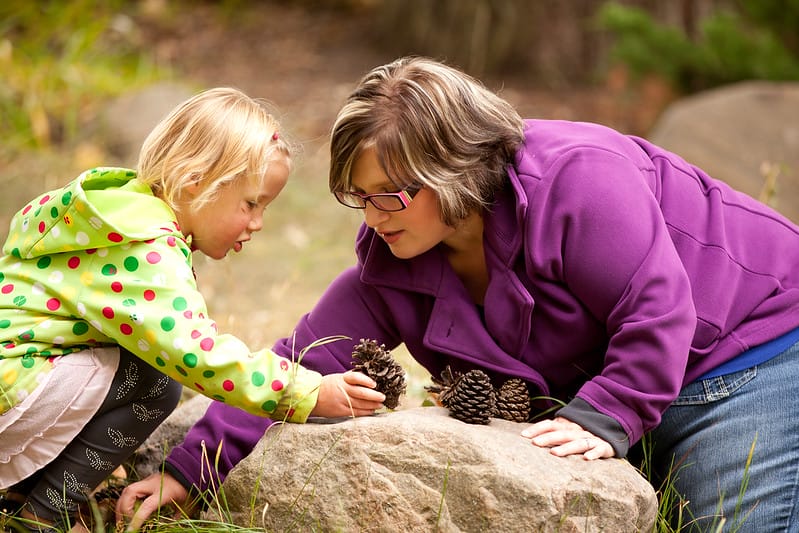 A woman and a young girl examine pine cones together on a rock outdoors, surrounded by trees.