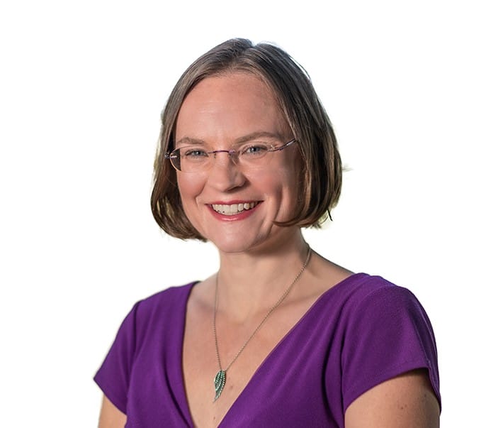 A smiling woman with short hair, wearing glasses and a purple top, isolated on a white background.
