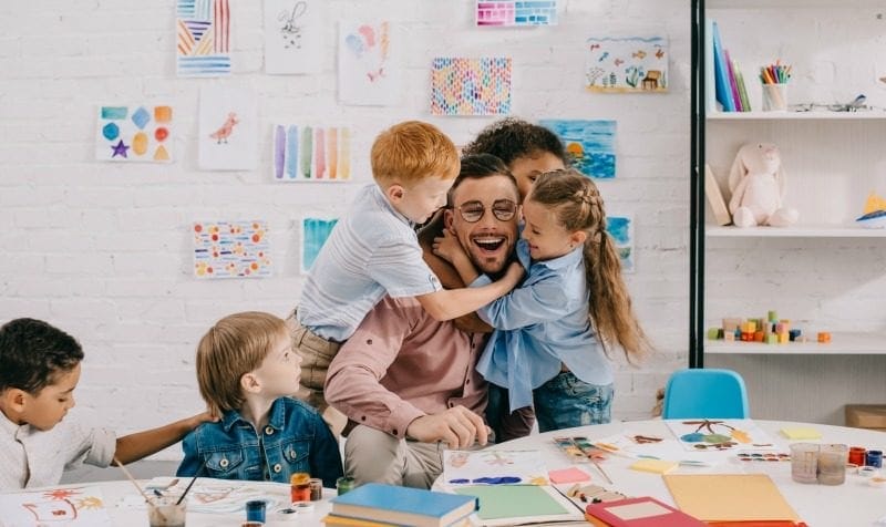 A joyful teacher surrounded by young children hugging him in a colorful classroom full of art supplies and drawings.