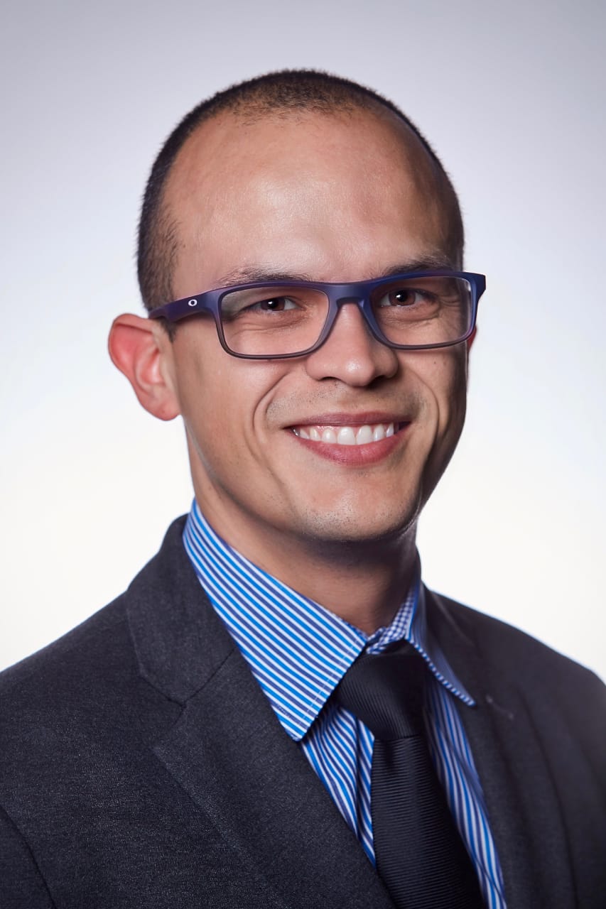 A professional portrait of a smiling bald man wearing glasses, a navy blazer, and a blue striped tie against a light background.