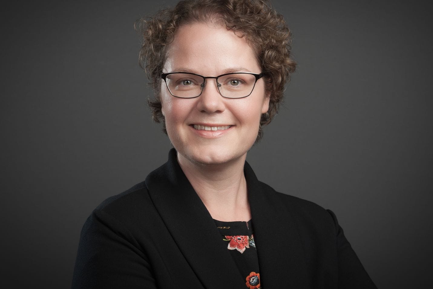 Professional headshot of a woman with curly hair wearing glasses, a black blazer, and a floral pin, smiling against a gray background.