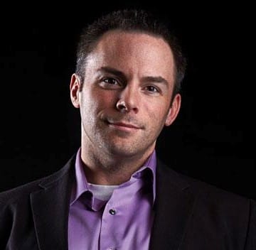 A man in a dark blazer and purple shirt, slightly smiling against a black background.