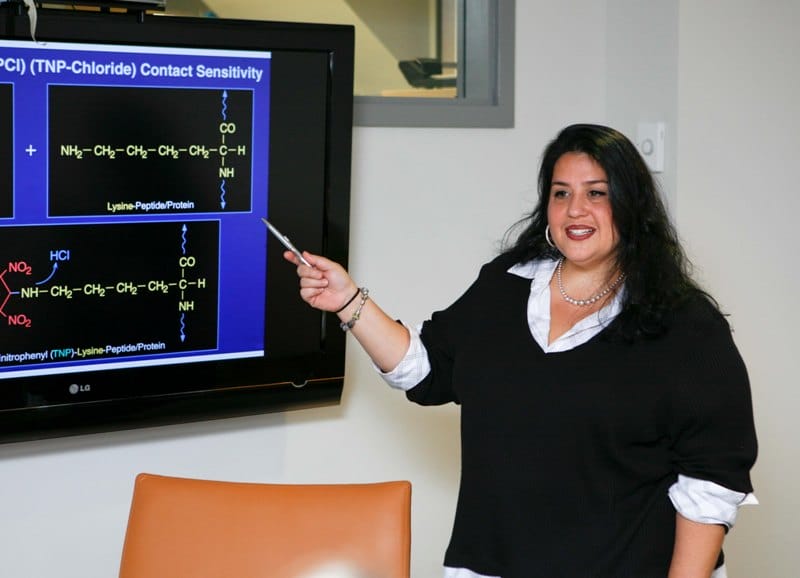 A woman presents a chemistry lecture, pointing at a complex molecular structure projected on a screen.