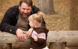 A man and a toddler girl with a ponytail laugh together over a wooden fence in a park.