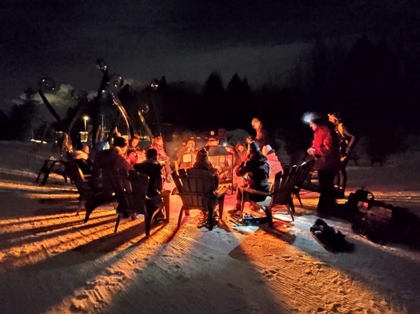 Group of people sitting around a fire pit on a snowy night, with skis and snowboards nearby, illuminated by firelight.